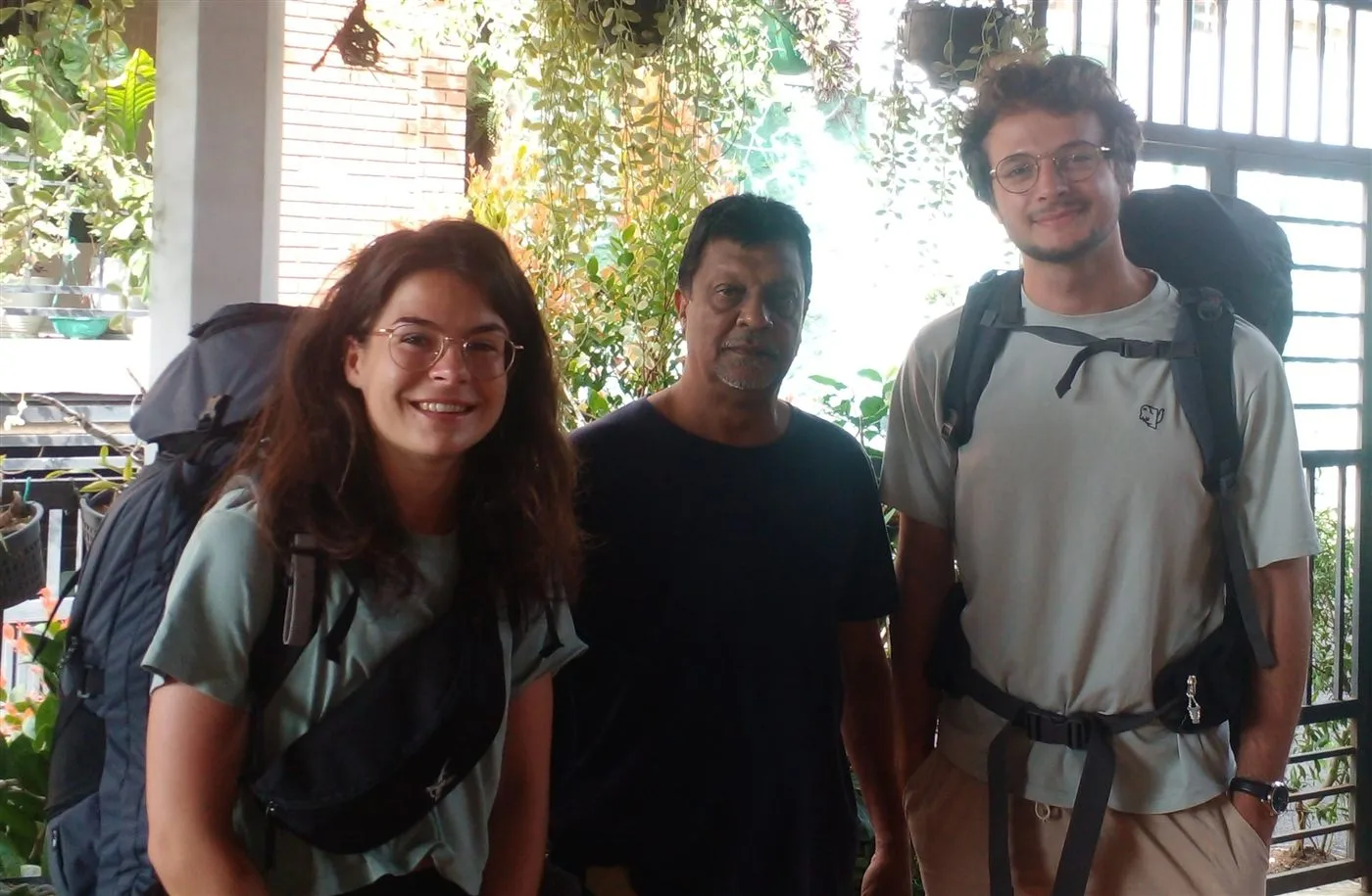 Two guests with backpacks standing with their host on the plant-filled veranda